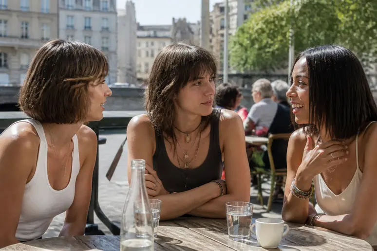 Trois jeunes femmes françaises discutent joyeusement dans un café ensoleillé, arborant différentes coupes mi-longues tendance et décontractées.